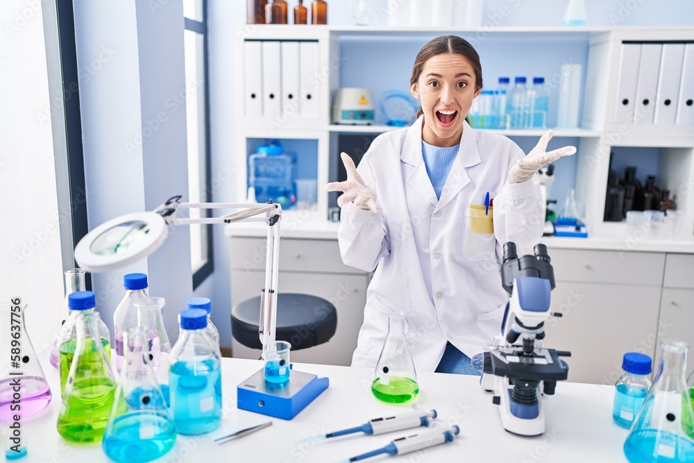 Fototapeta premium Young brunette woman working at scientist laboratory celebrating victory with happy smile and winner expression with raised hands