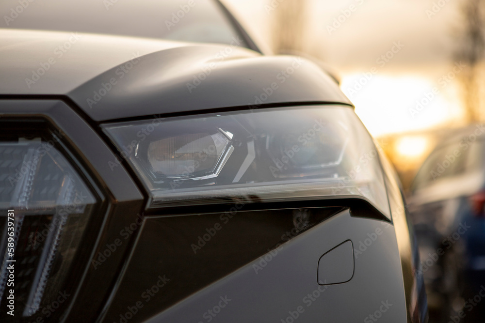 Left front full LED headlamp of modern black electric crossover SUV, dusk light in the background, blurred background, other cars behind. Part of grille, bonnet and bumper with lamp washer visible
