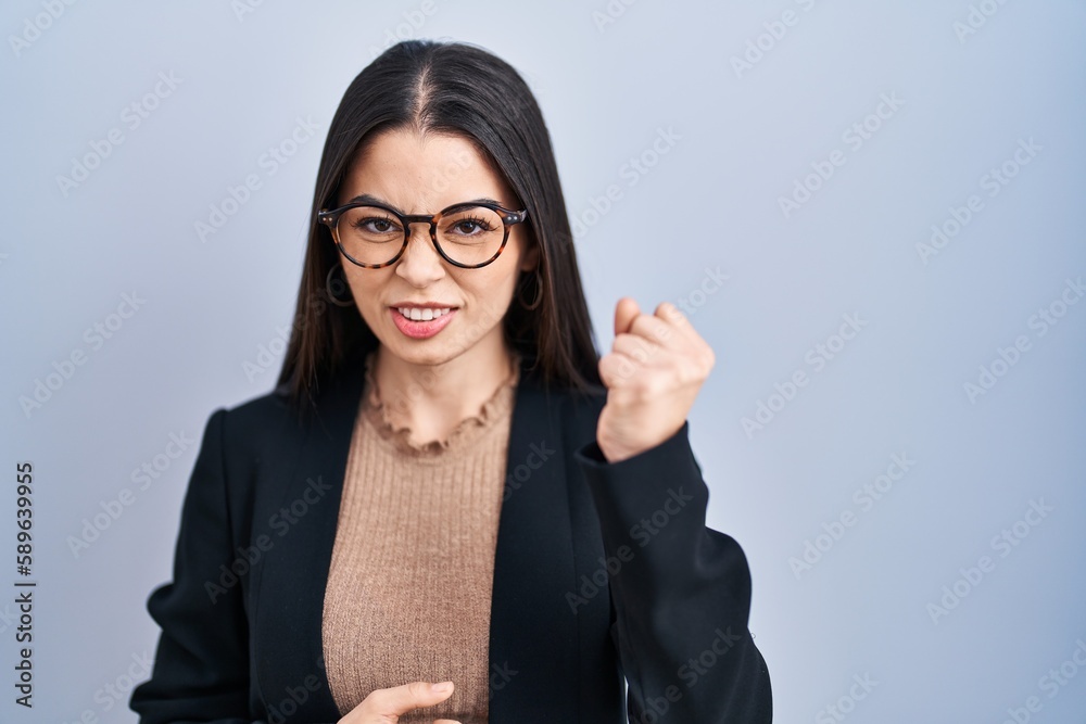 Young brunette woman standing over blue background angry and mad raising fist frustrated and furious while shouting with anger. rage and aggressive concept. Young brunette woman standing over blue background angry and mad raising fist frustrated and furious while shouting with anger. rage and aggressive concept.