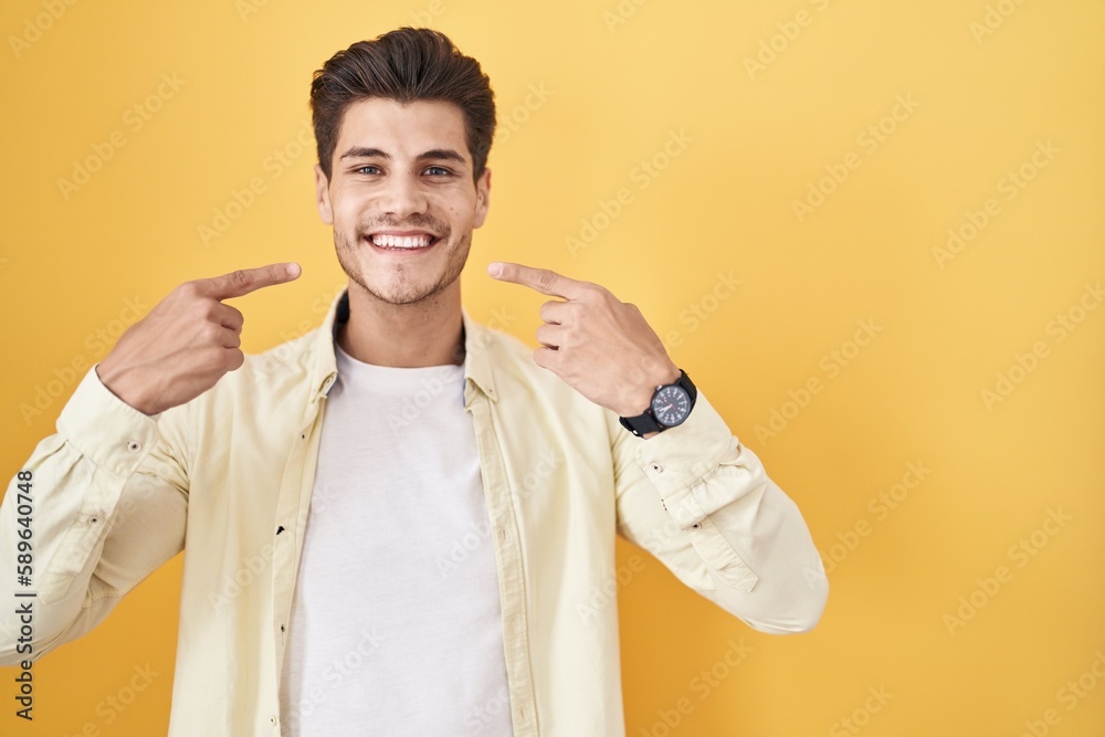 Young hispanic man standing over yellow background smiling cheerful showing and pointing with fingers teeth and mouth. dental health concept. Young hispanic man standing over yellow background smiling cheerful showing and pointing with fingers teeth and mouth. dental health concept.