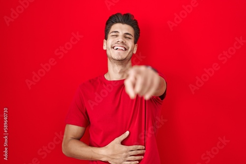 Young hispanic man standing over red background laughing at you, pointing finger to the camera with hand over body, shame expression
