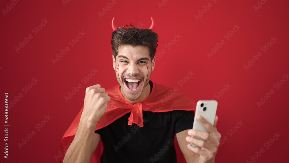 Young hispanic man wearing devil costume using smartphone over isolated red background Young hispanic man wearing devil costume using smartphone over isolated red background