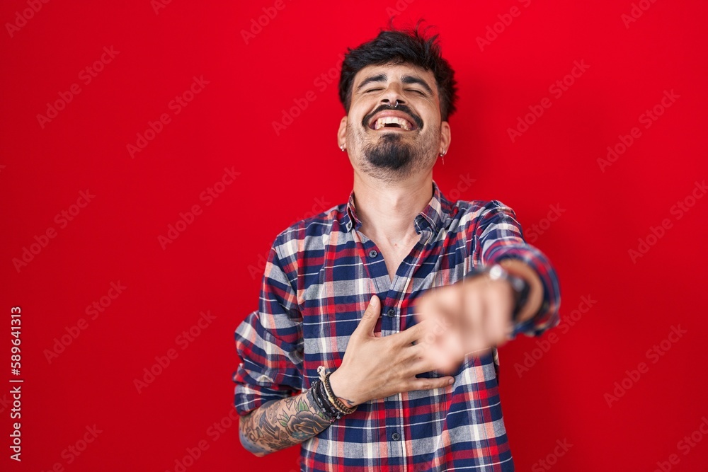 Young hispanic man with beard standing over red background laughing at you, pointing finger to the camera with hand over body, shame expression Young hispanic man with beard standing over red background laughing at you, pointing finger to the camera with hand over body, shame expression