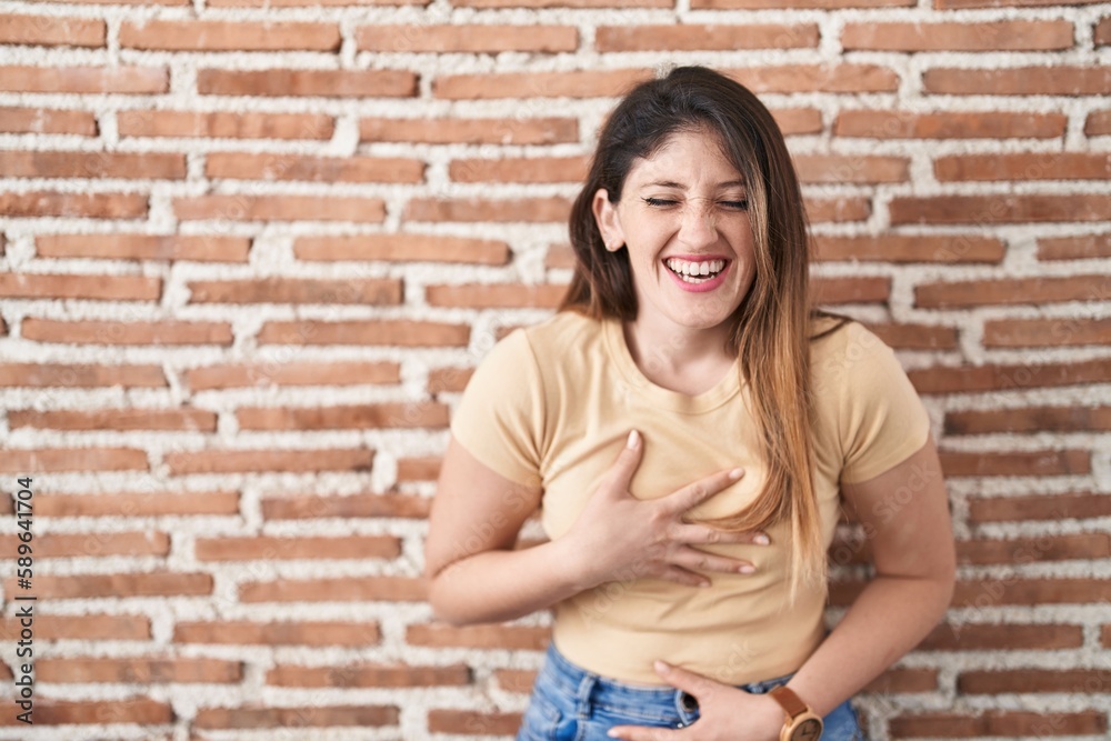 Young brunette woman standing over bricks wall smiling and laughing hard out loud because funny crazy joke with hands on body.