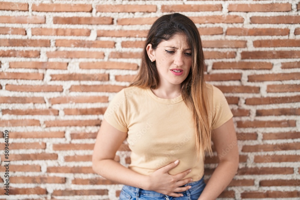 Young brunette woman standing over bricks wall with hand on stomach because indigestion, painful illness feeling unwell. ache concept. Young brunette woman standing over bricks wall with hand on stomach because indigestion, painful illness feeling unwell. ache concept.