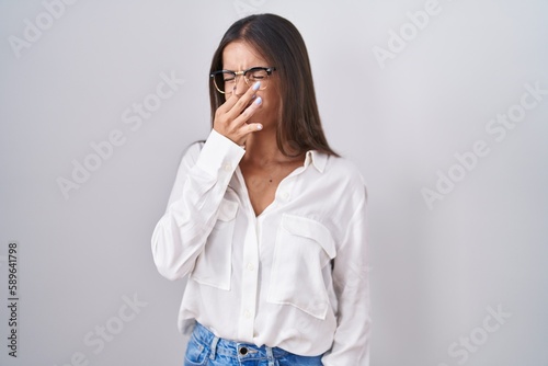 Young brunette woman wearing glasses smelling something stinky and disgusting, intolerable smell, holding breath with fingers on nose. bad smell