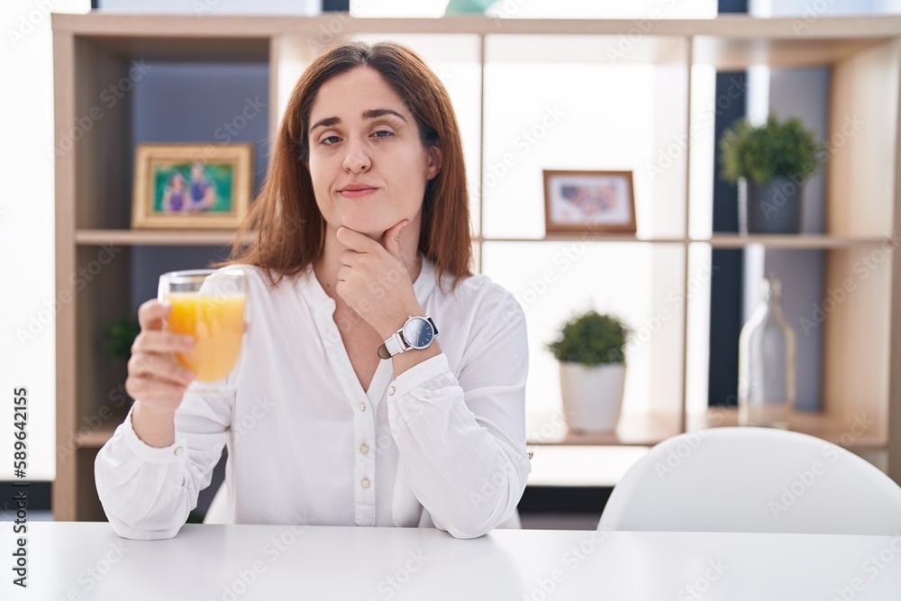 Brunette woman drinking glass of orange juice looking confident at the camera smiling with crossed arms and hand raised on chin. thinking positive. Brunette woman drinking glass of orange juice looking confident at the camera smiling with crossed arms and hand raised on chin. thinking positive.