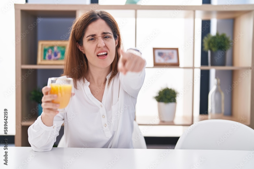 Brunette woman drinking glass of orange juice pointing displeased and frustrated to the camera, angry and furious with you Brunette woman drinking glass of orange juice pointing displeased and frustrated to the camera, angry and furious with you