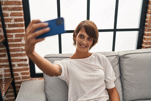 Young beautiful hispanic woman make selfie by smartphone sitting on sofa at home