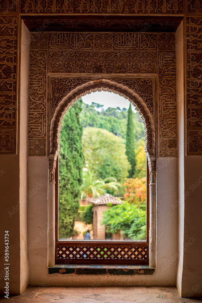 Window decorated with reliefs from the Nasrid palaces of the Alhambra in Granada, Spain with the blurred and colorful gardens in the background