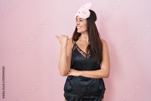 Young brunette woman wearing sleep mask and pyjama smiling with happy face looking and pointing to the side with thumb up.