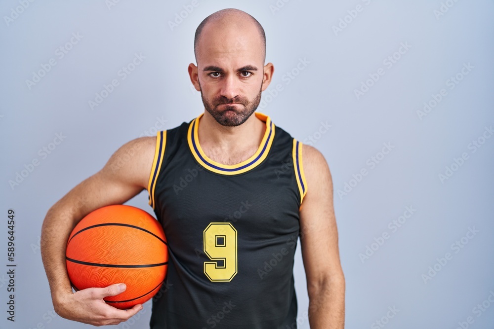 Young bald man with beard wearing basketball uniform holding ball skeptic and nervous, frowning upset because of problem. negative person.