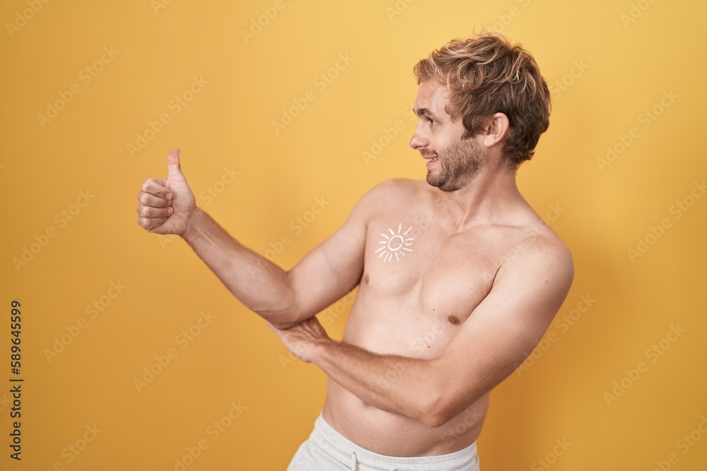 Caucasian man standing shirtless wearing sun screen looking proud, smiling doing thumbs up gesture to the side