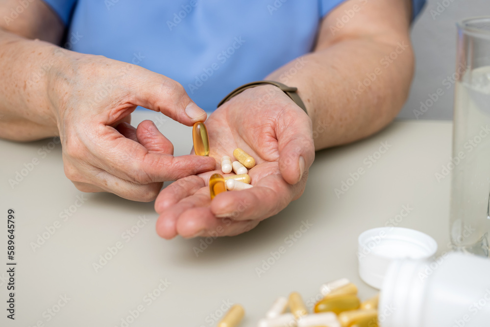 Senior woman with wrinkled old hands at the table holding Omega 3 capsule, vitamins and various pills for treatment. Healthcare and medicine concept