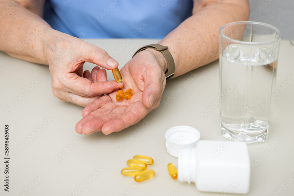 Senior woman with wrinkled old hands at the table holding omega 3 yellow capsules, fish oil pills, Healthcare and medicine concept Senior woman with wrinkled old hands at the table holding omega 3 yellow capsules, fish oil pills, Healthcare and medicine concept