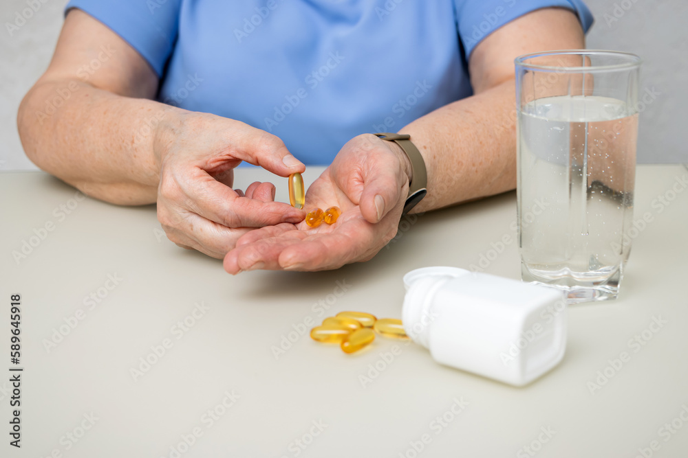 Senior woman with wrinkled old hands at the table holding omega 3 yellow capsules, fish oil pills, Healthcare and medicine concept Senior woman with wrinkled old hands at the table holding omega 3 yellow capsules, fish oil pills, Healthcare and medicine concept