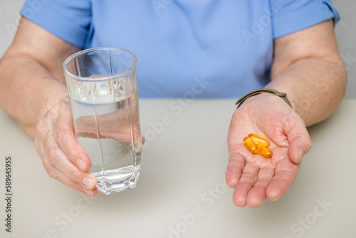 Senior woman with wrinkled old hands at the table holding omega 3 yellow capsules, fish oil pills and water glass. Healthcare and medicine concept