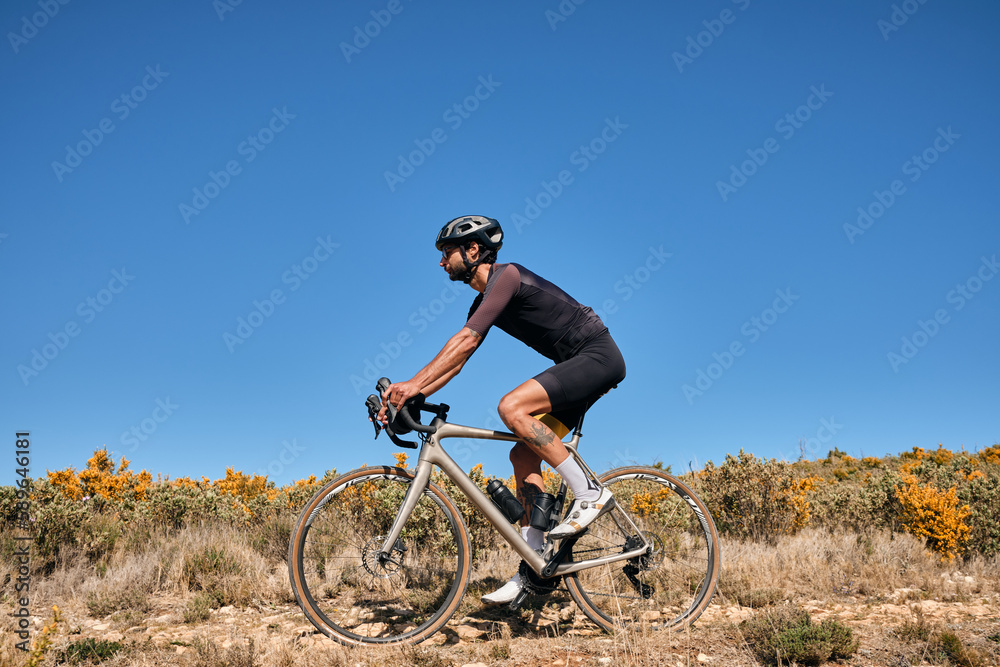 Fototapeta premium Man riding gravel bike on gravel road in mountains with scenic view.Cyclist riding on gravel bike.Cyclist practicing on gravel road.Man cyclist wearing cycling kit and helmet.Alicante region in Spain.