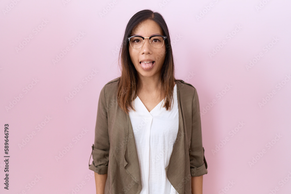 Middle age chinese woman wearing glasses over pink background sticking tongue out happy with funny expression. emotion concept. Middle age chinese woman wearing glasses over pink background sticking tongue out happy with funny expression. emotion concept.
