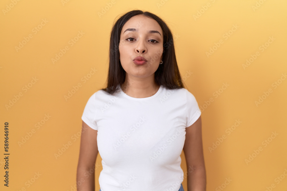 Young arab woman wearing casual white t shirt over yellow background looking at the camera blowing a kiss on air being lovely and sexy. love expression. Young arab woman wearing casual white t shirt over yellow background looking at the camera blowing a kiss on air being lovely and sexy. love expression.