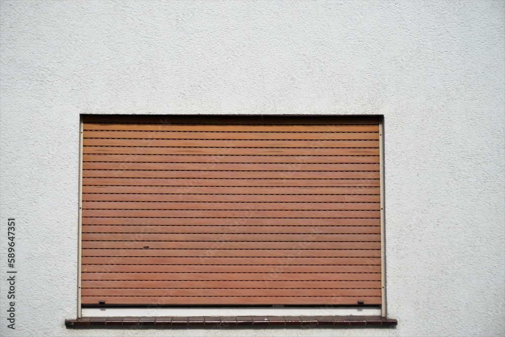 Fototapeta premium Weiße Hausfassade mit Fenster und runtergezogenen braun-roten alten Jalousien bei Sonne am Nachmittag im Frühling