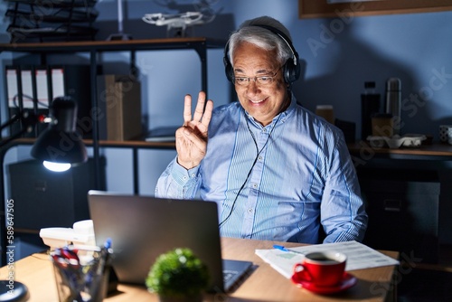 Hispanic senior man wearing call center agent headset at night showing and pointing up with fingers number three while smiling confident and happy.