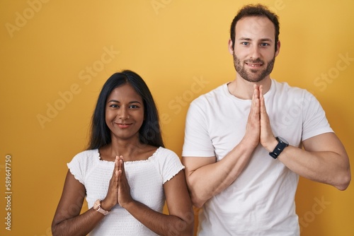 Interracial couple standing over yellow background praying with hands together asking for forgiveness smiling confident.