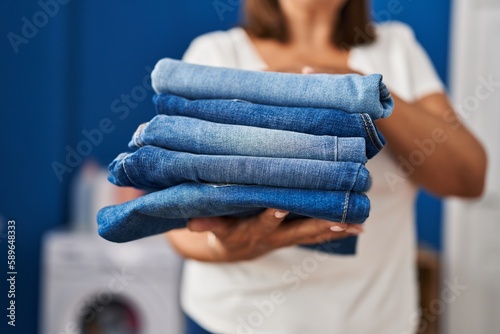 Middle age hispanic woman holding folded jeans at laundry room