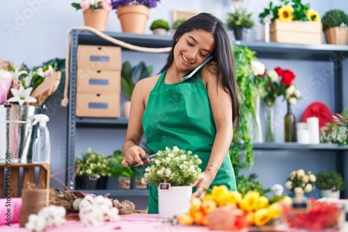 Young hispanic woman florist talking on smartphone cutting plant at florist shop