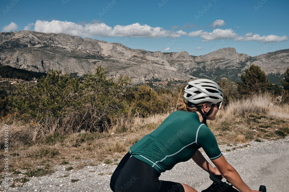 Close up back view of a female cyclist during ride.Woman cyclist wearing cycling kit and helmet riding on gravel bike.Cycling through stunning Spanish mountain landscapes. Close up back view of a female cyclist during ride.Woman cyclist wearing cycling kit and helmet riding on gravel bike.Cycling through stunning Spanish mountain landscapes.