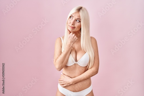 Caucasian woman wearing lingerie over pink background with hand on chin thinking about question, pensive expression. smiling with thoughtful face. doubt concept.