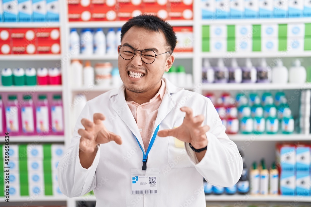 Chinese young man working at pharmacy drugstore smiling funny doing claw gesture as cat, aggressive and sexy expression Chinese young man working at pharmacy drugstore smiling funny doing claw gesture as cat, aggressive and sexy expression