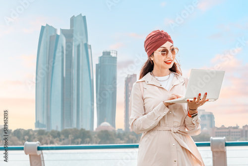 Photography A young woman takes advantage of the flexibility of remote work and education in Abu Dhabi, working on her laptop amidst the towering skyscrapers of the city's downtown