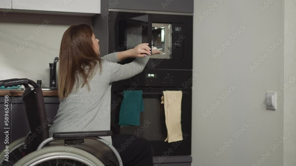 Woman in wheelchair takes out plate with heated food from microwave at home. Young mother smiles lovingly looking at entering kitchen toddler son Woman in wheelchair takes out plate with heated food from microwave at home. Young mother smiles lovingly looking at entering kitchen toddler son