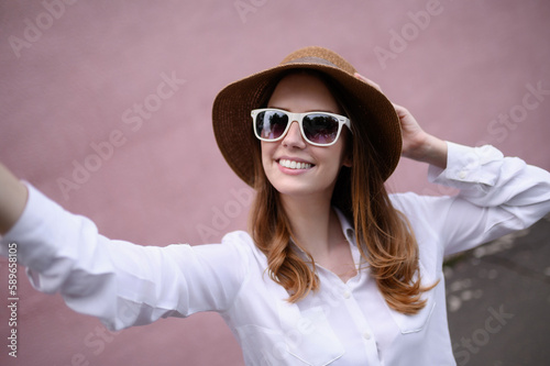 Young happy woman in summer clothes, sunglasses, straw hat posing on a pink background, taking a selfie portrait. Summer, technology concept © GRON777
