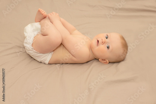 a baby with gray eyes in a diaper is lying in a crib, holding his legs with his hands and looking at the camera.