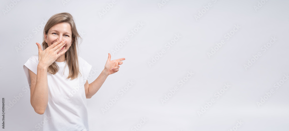 Laughing blonde woman on gray background pointing side and laughing covering her face. Long horizontal background. White clothe t-shirt and solid gray background Laughing blonde woman on gray background pointing side and laughing covering her face. Long horizontal background. White clothe t-shirt and solid gray background