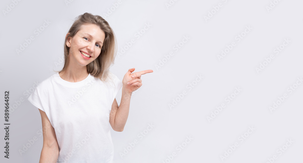 Attractive blonde woman pointing finger with calm relaxed smile. Long horizontal background. White clothe t-shirt and solid gray background Attractive blonde woman pointing finger with calm relaxed smile. Long horizontal background. White clothe t-shirt and solid gray background