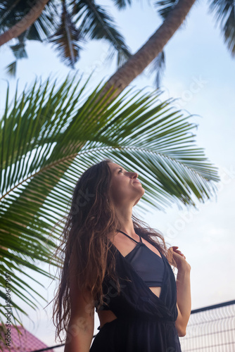 Pensive pretty young smiling woman in black dress on seacoast looking up at tropical background  summer vacation. Lovely beautiful lady on resting  travel lifestyle. Tropic holiday concept. Copy space