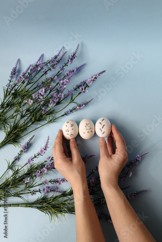 Easter, hands hold three white chicken painted eggs with a bunny on a blue background and with a bouquet of violet flowers. Easter holiday, minimalism, spring composition. Free copy space.