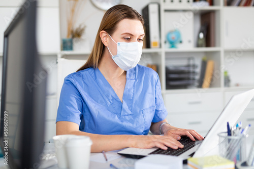 Portrait of young female doctor in face mask working on laptop in clinic office