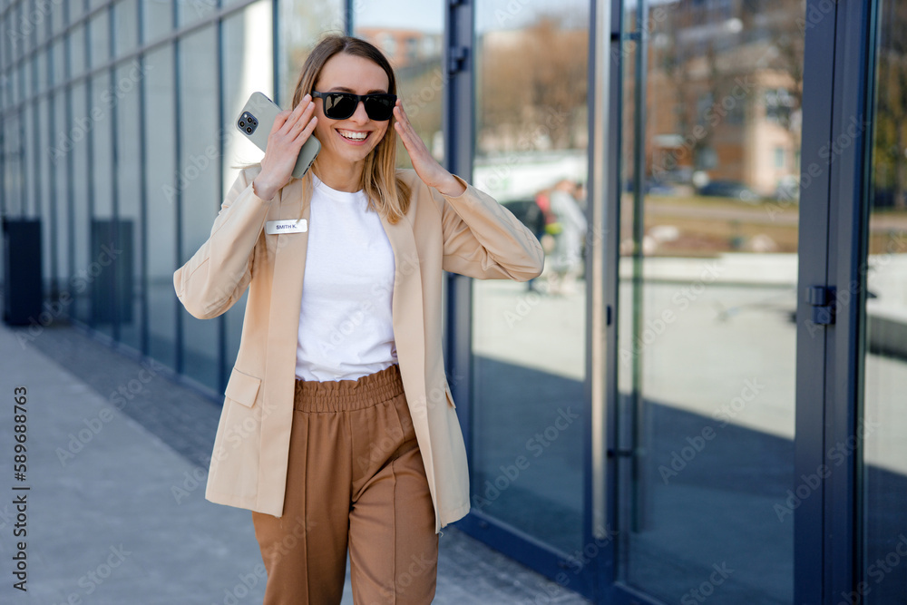 Charming manager smiling and looking at camera while walking near office center with crossed arms. Beautiful business woman wearing stylish formal jacket and trendy sunglasses
