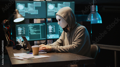 Cyber scammer wearing mask and hood to hack computer system, breaking into servers to steal big data. Masked man looking dangerous and scary, impostor creating security malware. Handheld shot. photo