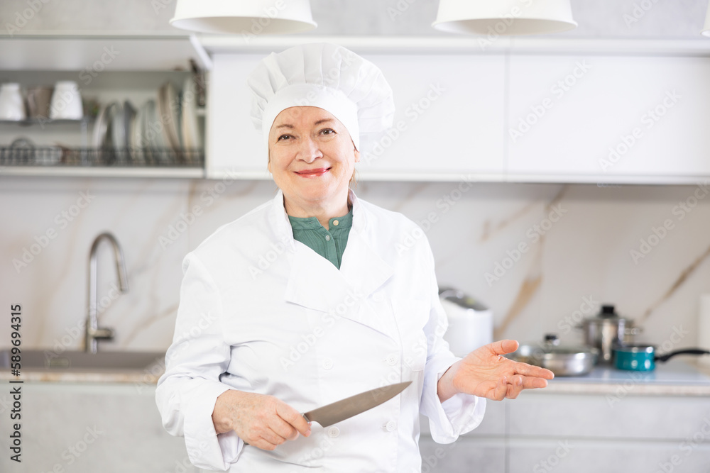 Portrait of a positive mature female chef standing with a knife in her hands in a home kitchen