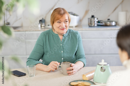 Mature woman sitting at a table in a home kitchen with a female guest, drinking tea together and discussing something