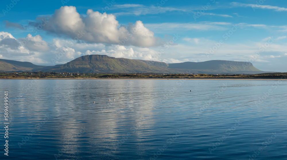 Panorama image on Benbulben mountains and blue water surface of Atlantic ocean with reflection of blue cloudy sky. Irish nature. County Sligo, Ireland. Popular tourist attraction. Stunning scenery.