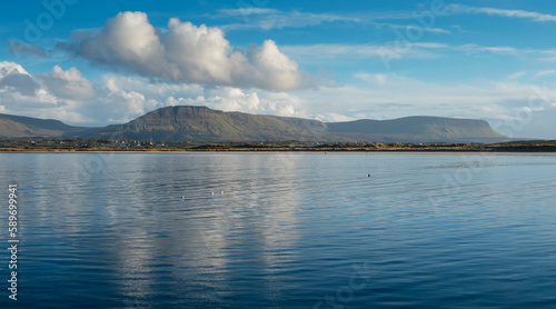 Panorama image on Benbulben mountains and blue water surface of Atlantic ocean with reflection of blue cloudy sky. Irish nature. County Sligo, Ireland. Popular tourist attraction. Stunning scenery.