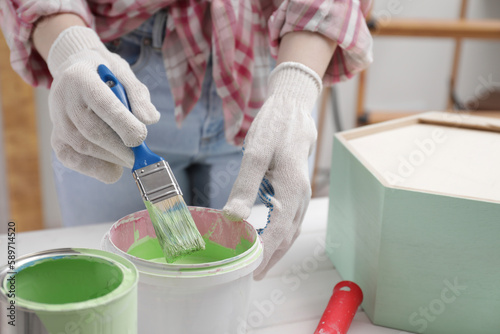 Woman dipping brush into bucket of green paint at white table indoors, closeup © New Africa