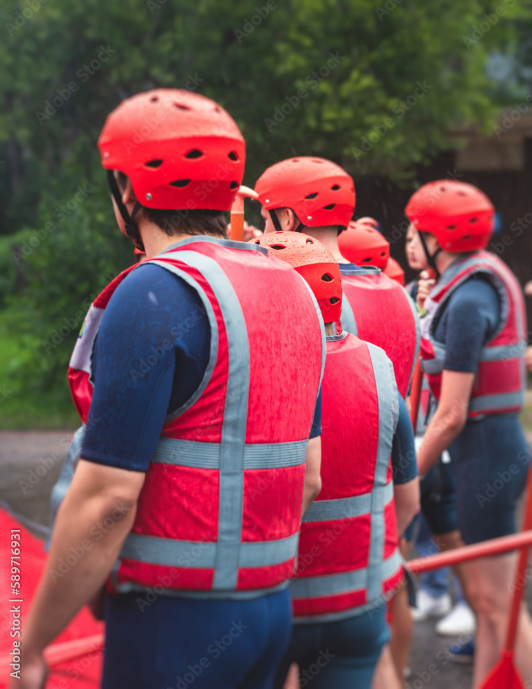 Obraz premium Group of sportsmen in wetsuits with paddles in helmets and life jackets vest prepare to extreme water sports, team before rafting, kayaking, canoeing in a summer day, equipment view with raft boat