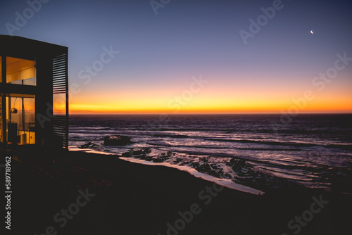 Oceanic tranquility: silhouette of house in the cliff with warm lights and white crescent moon illuminating the dark purple ocean, as waves gently crash onto the beach sand, under a serene sunset sky